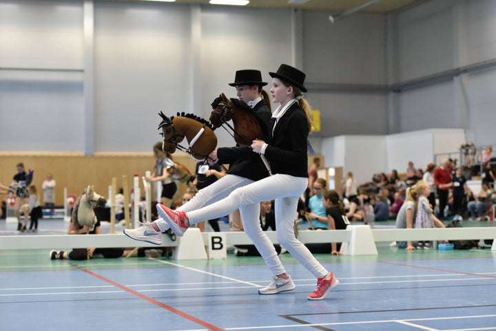 Dve deti vo formálnych odevoch podľa dress code pre Hobby Horsing súťažia v hale s Hobby Horses