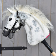 Hobby Horse LarDen "Hobby Horse Aspen with Black Halter", veľkosť S, s bielou plyšovou hlavou zdobenou čiernymi škvrnami, huňatou hrivou a čiernou ohlávkou, nasadený na drevenej tyči a vystavený pred sivou drevenou stenou.