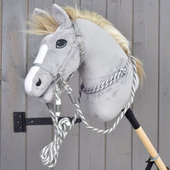 Set Start strieborný (ohlávka + nákrčník + vodidlo + opraty) od Hobby Horse LarDen na sivom plyšovom Hobby Horse s bielym pruhom na hlave, nasadenom na drevenej tyči a opretom o sivú drevenú stenu.