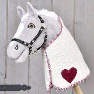 Plyšový Hobby Horse od Hobby Horse LarDen s bielosivou tvárou, čiernou uzdečkou a fleecovou Dekou Cube Red s červenou nášivkou v tvare srdca, nasadený na drevenej tyči na pozadí svetlého dreva.