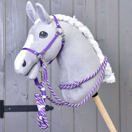 Plyšový Hobby Horse so sivou hlavou, bielou hrivou a bielou lysinou má sadu Set Start Fialový (ohlávka + nákrčník + vodidlo + opraty) od Hobby Horse LarDen a je upevnený na drevenej tyči pred sivou drevenou stenou.