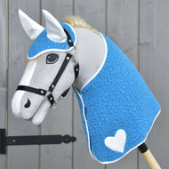 Hobby Horse LarDen Set Mystery zobrazuje sivého koňa s modrým textúrovaným poťahom pripomínajúcim deku, bielymi ušami, bielym srdcom na boku, čiernou uzdečkou, hustou bielou hrivou, nasadeného na tyči pred dreveným pozadím.