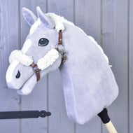 Hnedá ohlávka z koženky s kompletným barančekom od Hobby Horse LarDen na plyšovom svetlosivom Hobby Horse s bielou hrivou a odznakmi, nasadenom na drevenej tyči a odfotografovanom pred pozadím zo sivých drevených panelov.