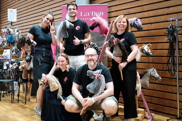 Päť usmievavých dospelých pózuje vnútri so svojimi Hobby Horse a oslavuje Hobby Horsing. Štyria stoja a jeden kľačí; všetci majú na sebe čierne tričká. Za nimi je drevom obložená stena a ružový nápis „LarDen“, s vystavenými rôznymi Hobby Horse.