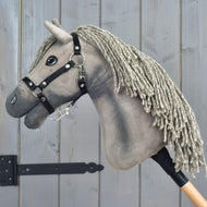 Hobby Horse Swing s čiernou ohlávkou - veľkosť S od Hobby Horse LarDen, s čiernou ohlávkou, inšpirovaný eleganciou hannoverského žrebca, so sivou priadzovou hrivou, čiernou uzdečkou a gombíkovými očami, na drevenej tyči, odfotografovaný pred sivým dreveným pozadím.