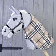 Hobby Horse LarDen „Deka Cremo England“ na sivom Hobby Horse s čiernou ohlávkou a béžovým károvaným kabátikom, doplnená čabrakou na ochranu pred hmyzom, nasadenom na drevenej tyči pred sivou drevenou stenou.