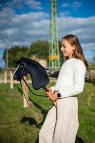 Dievča v bielom svetri stojí na slnečnej lúke a drží Hobby Horse Loki with Black Halter od Hobby Horse LarDen s čiernou plyšovou hlavou, bielou lysinou a čiernou priadzovou hrivou, s kompletnou uzdečkou Pure Black bez nánosníka