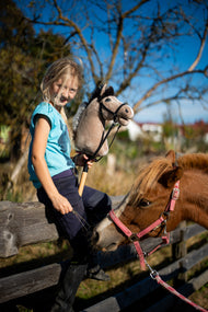 Blondínka v tyrkysovom tope sedí na drevenej ohrade výbehu s Hobby Horse Piccolino s čiernou ohlávkou od Hobby Horse LarDen, svetlobéžovou plyšovou hlavou s tmavou hrivou z priadze, za ňou ryšavý poník, s kompletnou čiernou uzdečkou z koženky