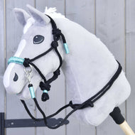 Povrazový set Turquoise od Hobby Horse LarDen je zobrazený na sivom plyšovom Hobby Horse s bielou hrivou, čiernou uzdečkou a tyrkysovými detailmi, upevnenom na drevenej tyči pred sivou drevenou stenou.