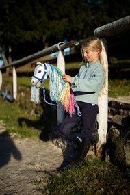 Dievča v mätovej mikine pri bielom zábradlí jazdiarne drží bieleho Hobby Horse s dúhovou hrivou na modro pruhovanom vodidle