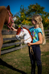 Dievča v tyrkysovom tričku drží Hobby Horse Rufus s čiernou ohlávkou od Hobby Horse LarDen, bielu plyšovú hlavu so škvrnami Appaloosa a červenou hrivou z priadze, vedľa ryšavého poníka na slnečnom výbehu, s bordovým setom z koženky – uzdečkou, opratami a martingalom