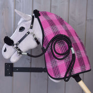 Sivý Hobby Horse zo setu „Set Harmony Pink“ od Hobby Horse LarDen je zavesený na drevenej stene a má na sebe ružovo-čiernu károvanú stajňovú deku, čiernu ohlávku a vedľa seba ladiace stočené vodidlo.