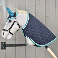 Set Supreme Turquoise od Hobby Horse LarDen so sivým plyšovým Hobby Horse s modrými ušami, čiernou uzdečkou a prešívanou tmavomodrou dekou s tyrkysovým lemom, nasadený na drevenej tyči pred stenou zo svetlého dreva.