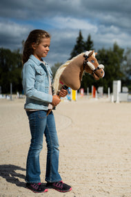 Dievča v džínsovej bunde stojí na pieskovom kolbisku a drží Hobby Horse Smart with Black Halter od LarDen, za ním sú skokové tyče – palominová plyšová hlava s dlhou krémovou priadzovou hrivou – s čiernou koženkovou ohlávkou s úplným kožušinovým lemom