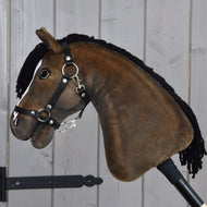 Hobby Horse Sundance s čiernou ohlávkou od Hobby Horse LarDen s plyšovým prevedením, čiernou priadzovou hrivou, uzdečkou a oprátami, zachytávajúci temperamentný vzhľad anglického plnokrvného žrebca, upevnený na tyči na pozadí svetlej steny z drevených dosiek.