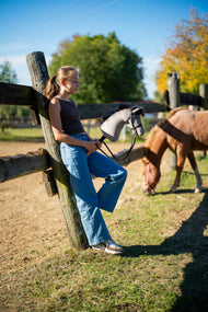 Dievča opreté o ohradu výbehu na jesennom slnku s Hobby Horse Thor v čiernej uzdečke, za ňou sa pasie ryšavý poník