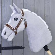 Ohlávka s barančekom z koženky Walnut od Hobby Horse LarDen na sivom plyšovom Hobby Horse s bielou hrivou, hnedou koženkovou uzdečkou a veľkými čiernymi očami na drevenej tyči, opretom o sivú drevenú stenu.