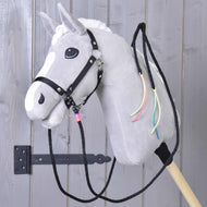 Hobby Horse LarDen "Westernové opraty Texas delené", sivý plyšový koník s bielou hrivou, čiernou uzdečkou, delenými westernovými oprátami a farebnými stuhami na krku, nasadený na drevenej tyči pred sivými drevenými dverami.