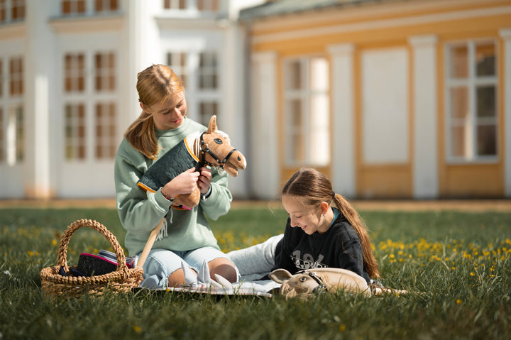 Dievča vo svetlozelenej mikine sedí na tráve vedľa prúteného koša s výbavou pre Hobby Horse a jemne drží svetlohnedého Hobby Horse s čiernou uzdečkou, bielou hrivou a prešívaným prehozom. Ďalšie dieťa je čiastočne vidieť na deke neďaleko.