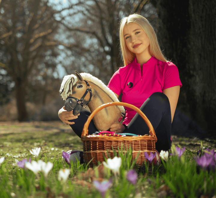 Mladá žena v jasnoružovom tričku sedí vonku na tráve, drží hračku Hobby Horse a prútený košík plný farebných vajec, obklopená rozkvitnutými bielymi a fialovými kvetmi.