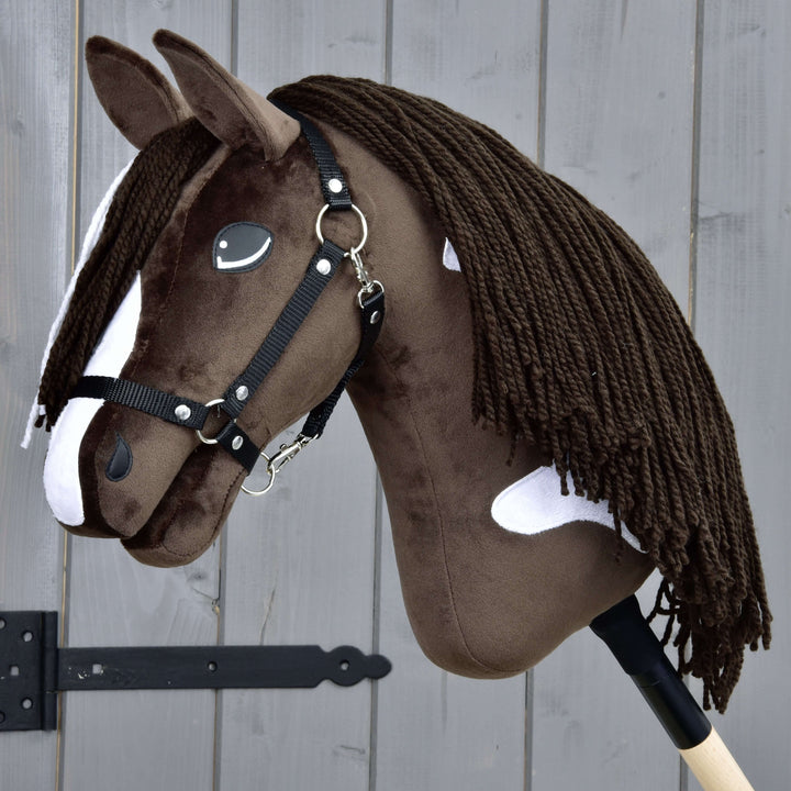 Realistický hnedobiely Hobby Horse s čiernou uzdečkou a dlhou hrivou z priadze, nasadený na drevenej tyči, umiestnený pred sivým dreveným plotom.