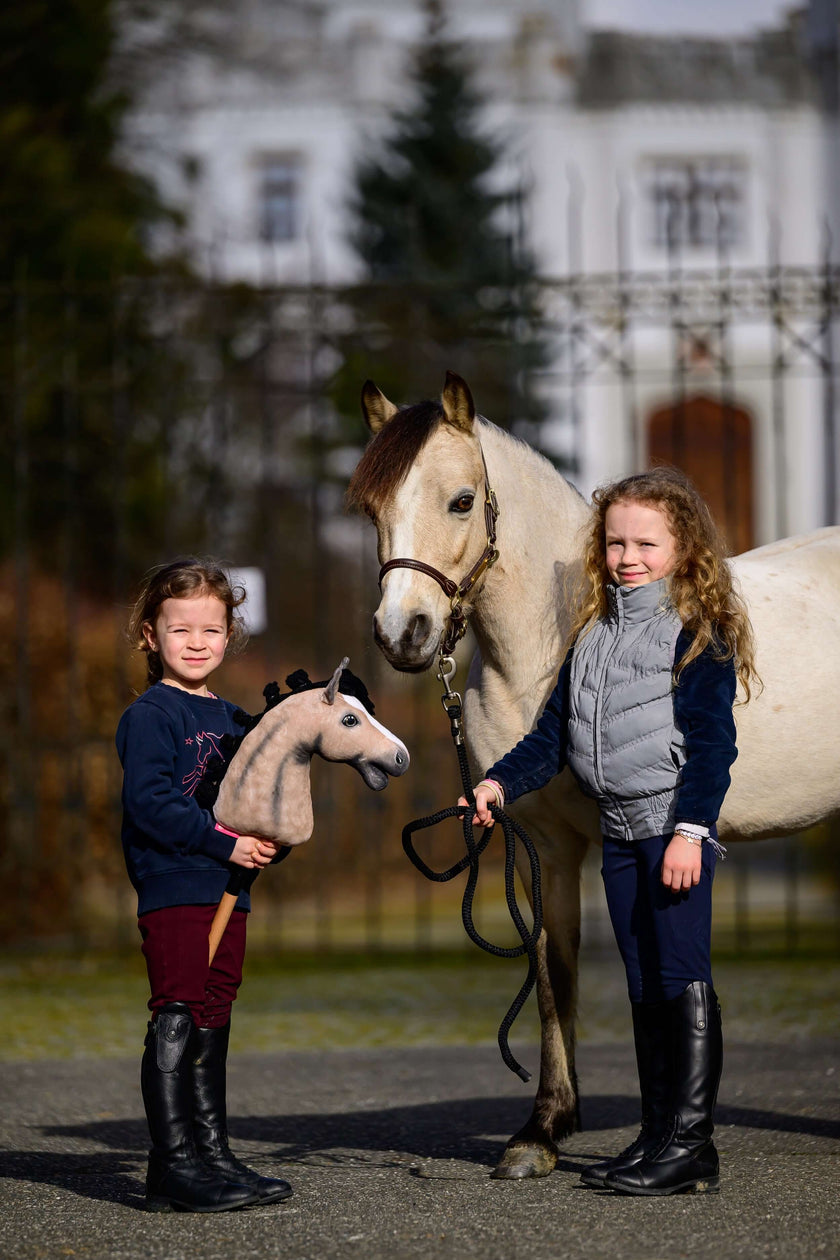 Dve malé dievčatá stoja vonku vedľa bieleho koňa; jedna drží hračku Hobby Horse ako odraz ich lásky k Hobby Horsingu, zatiaľ čo druhá drží opraty skutočného koňa. V pozadí vidno rozmazanú budovu a stromy.