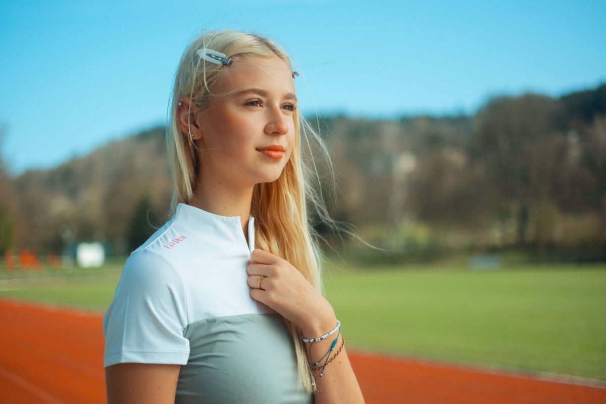 Mladá blond žena v športovom oblečení stojí na vonkajšej bežeckej dráhe a zamyslene drží svoj Hobby Horse. V pozadí je zelená tráva, stromy a modrá obloha, čo vystihuje jej vášeň pre Hobby Horsing.