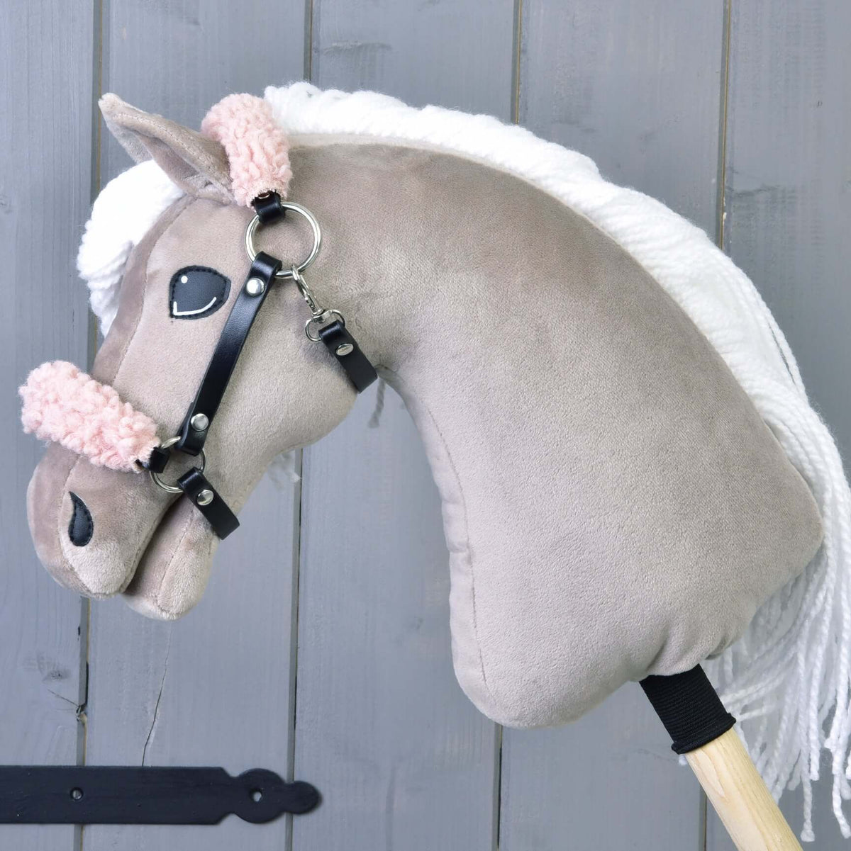Čierna ohlávka z koženky so staroružovým barančekom od Hobby Horse LarDen na plyšovom Hobby Horse so svetlohnedou hlavou, bielou hrivou, ružovou chlpatou uzdečkou a čiernou koženkovou ohlávkou na drevenej tyči pred sivou drevenou stenou.