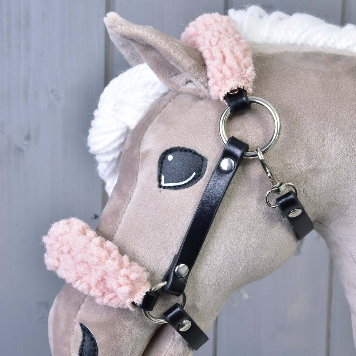 Detail hlavy plyšového koňa s bielou priadzovou hrivou, čiernym plsteným okom a čiernou koženkovou ohlávkou Hobby Horse LarDen so staroružovým barančekom na svetlosivom drevenom pozadí.