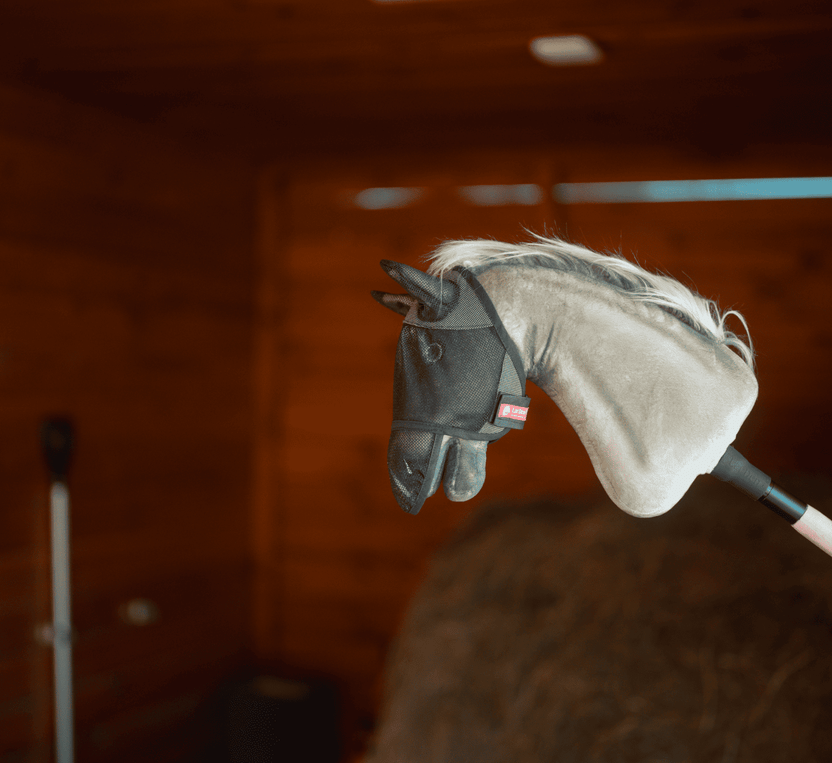 Plyšová hlava Hobby Horse na tyči s čiernou sieťovanou maskou proti hmyzu je odfotená v interiéri s drevenými stenami a balíkom sena – ideálna na nápaditú zábavu s Hobby Horsingom.