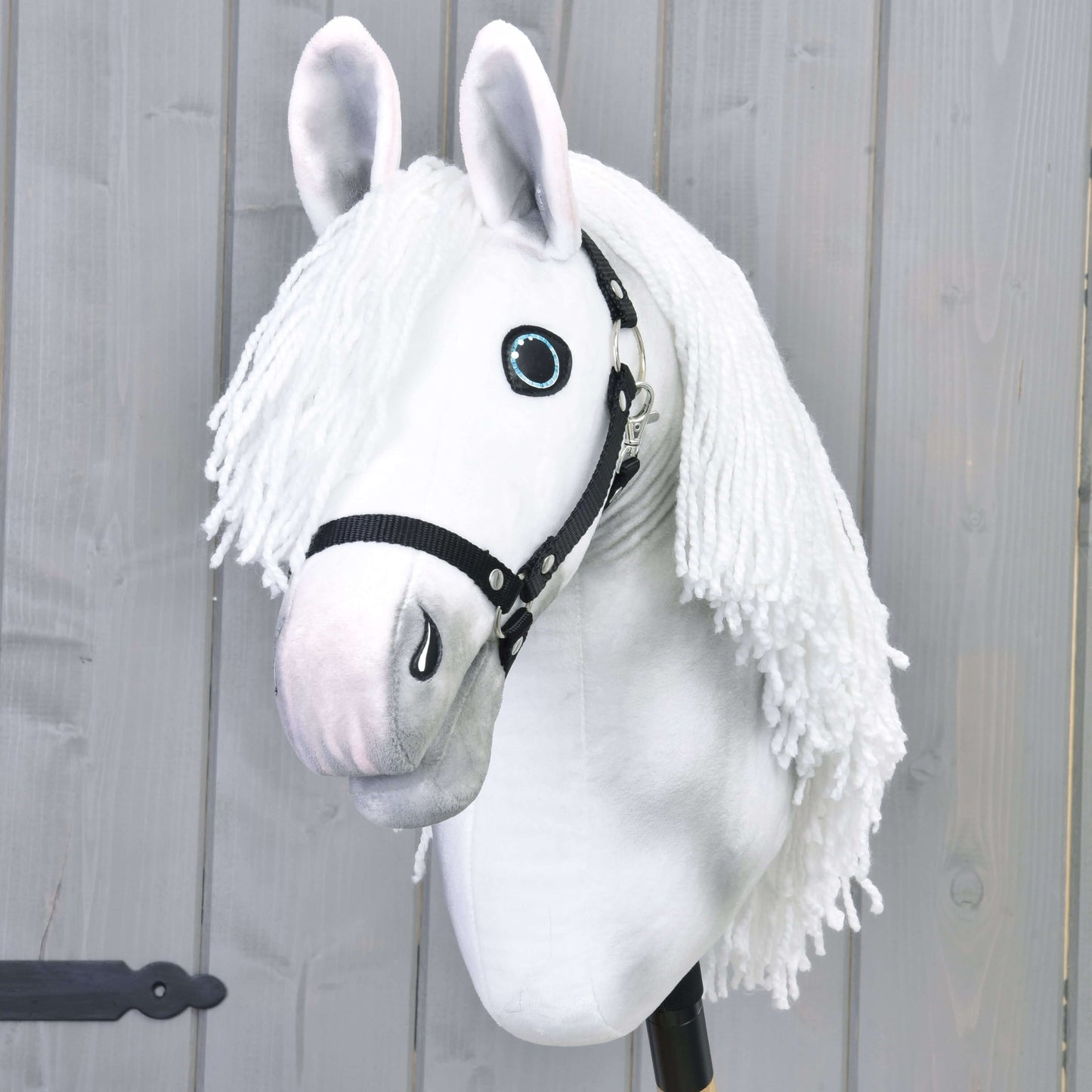 Hobby Horse Elektra s čiernou ohlávkou od Hobby Horse LarDen s ručne maľovanou bielou kobylou, dlhou priadzovou hrivou, modrými očami a čiernou ohlávkou, nasadený na tyči pred svetlosivou drevenou stenou.