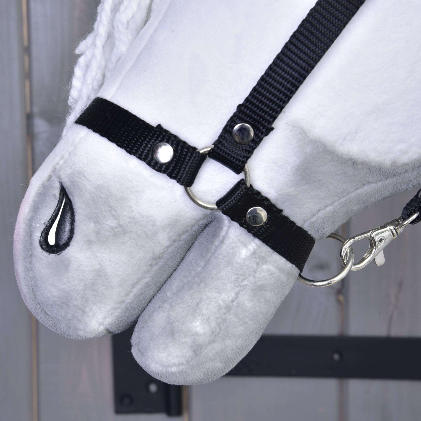 Detail Hobby Horse Elektra s čiernou ohlávkou od Hobby Horse LarDen s plyšovou bielou tvárou, čiernou látkovou ohlávkou so striebornými krúžkami a karabínou a čierne prešitou nozdrou, na drevenom pozadí.