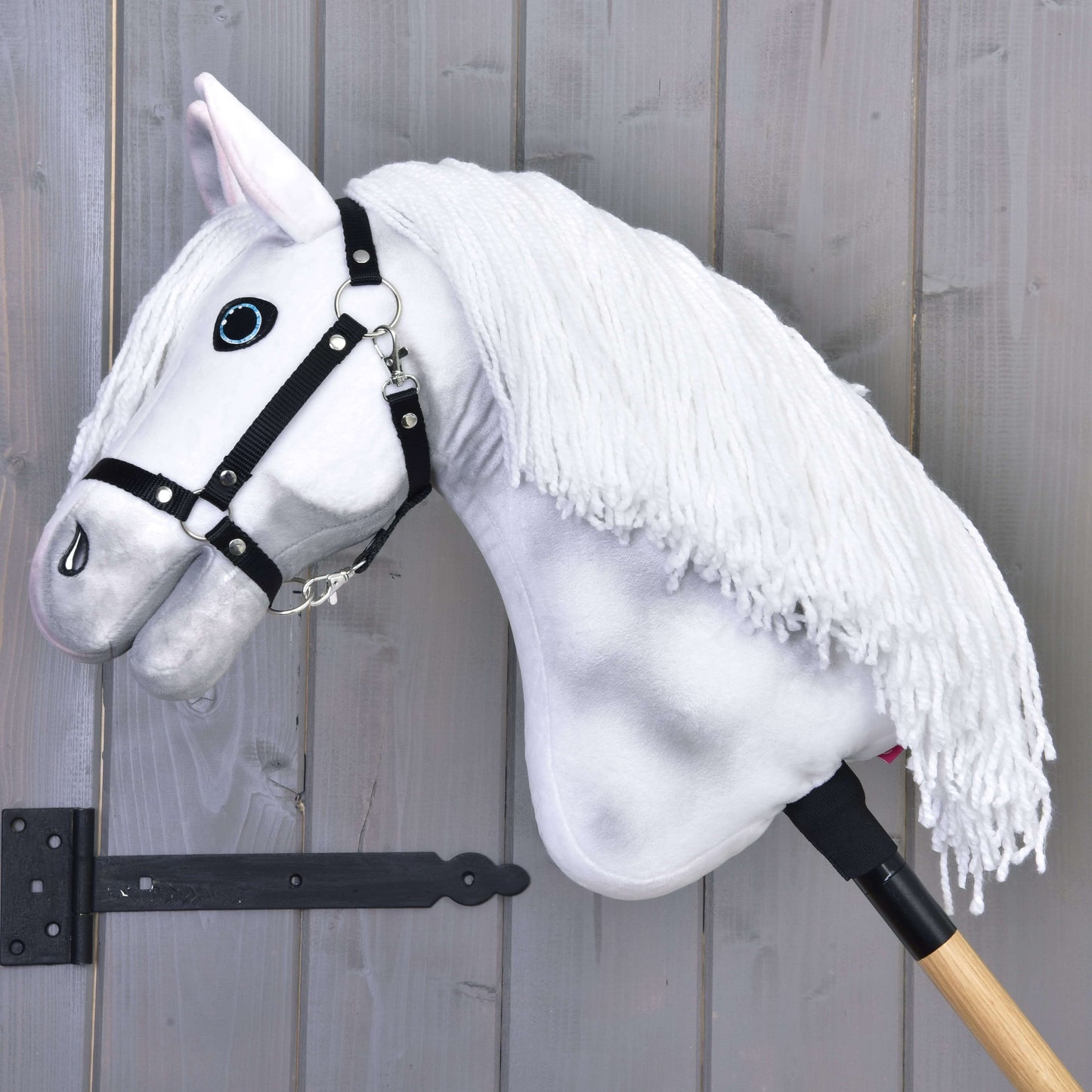 Hobby Horse Elektra s čiernou ohlávkou od Hobby Horse LarDen s ručne maľovanou bielou hrivou, nasadený na drevenej tyči a opretý o sivú drevenú stenu.