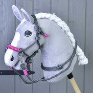Plyšový šedo-bílý Hobby Horse se zapletenou hřívou a černýma očima předvádí Provazový set Šedá (vel. M) od Hobby Horse LarDen před dřevěným obložením a zvýrazňuje jedinečnou ručně vyráběnou jezdeckou výbavu.