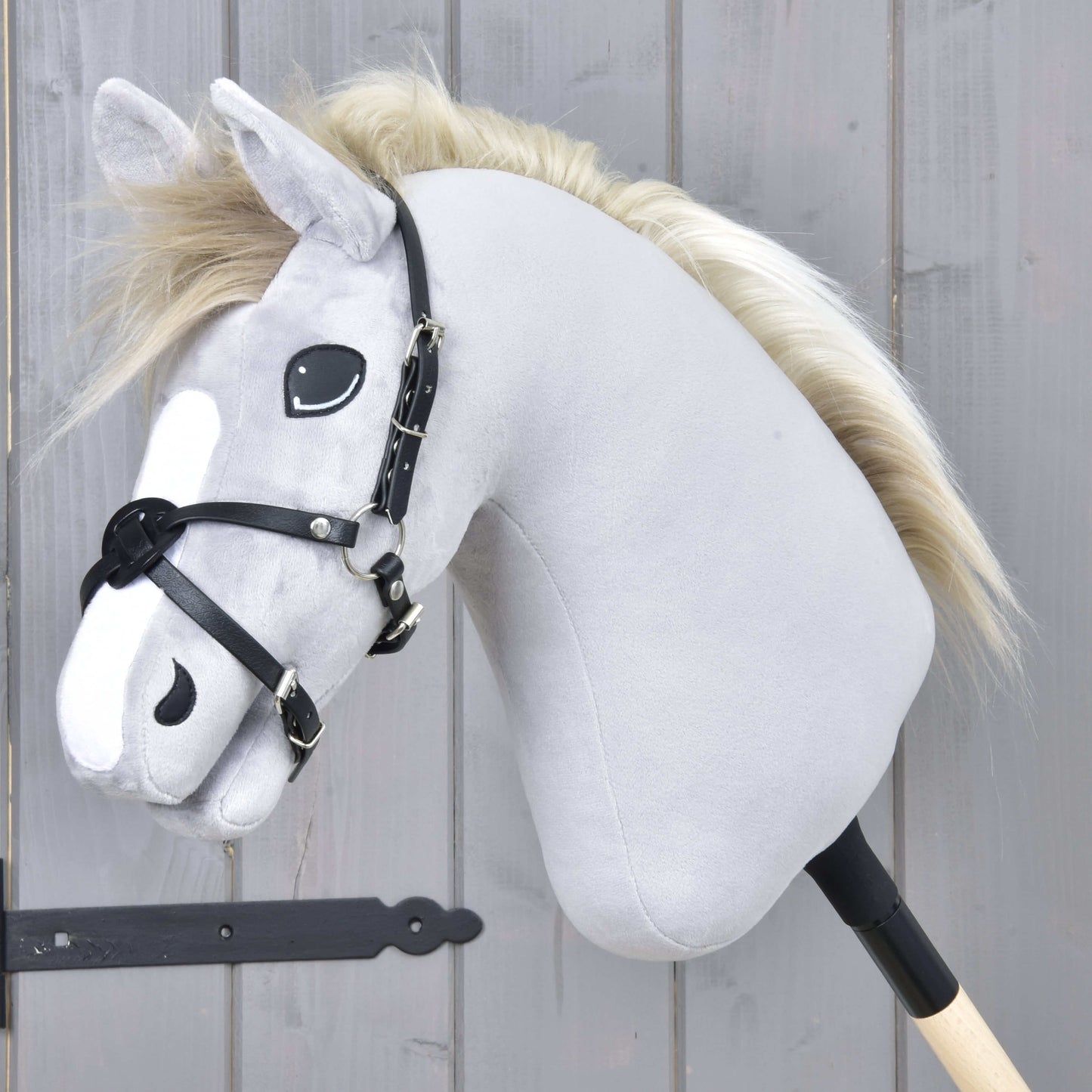 Mexický nánosník černý od Hobby Horse LarDen na šedém plyšovém Hobby Horse s černou uzdečkou, nánosníkem z koženky a světlou hřívou, na tyči před šedou dřevěnou stěnou.