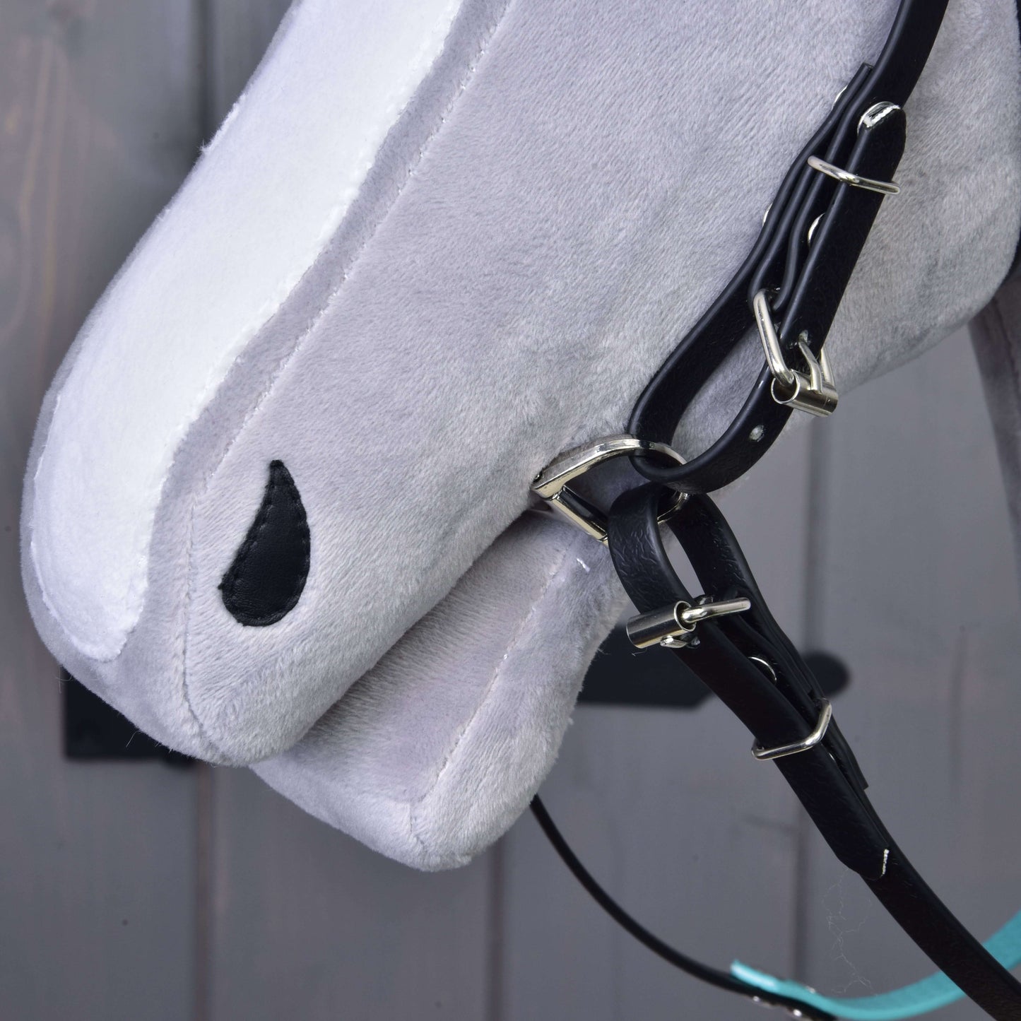 Detail sivého Hobby Horse od Hobby Horse LarDen s Kompletnou uzdečkou s tyrkysovou čelenkou bez nánosníka a opratami, s čiernym okom v tvare kvapky, na rozmazanom sivom pozadí.