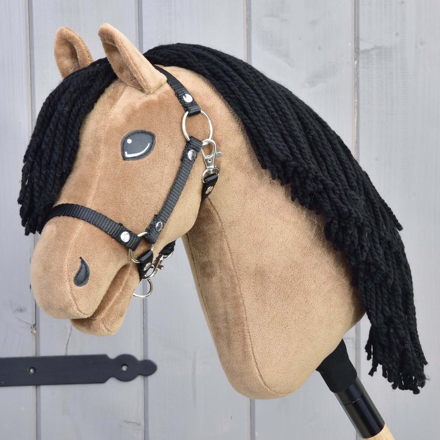 Hobby Horse Samantha s čiernou ohlávkou od Hobby Horse LarDen má béžovú látkovú hlavu, čiernu priadzovú hrivu a je inšpirovaná kobylou anglického plnokrvníka; je zobrazená na drevenej tyči pred sivými drevenými panelmi.