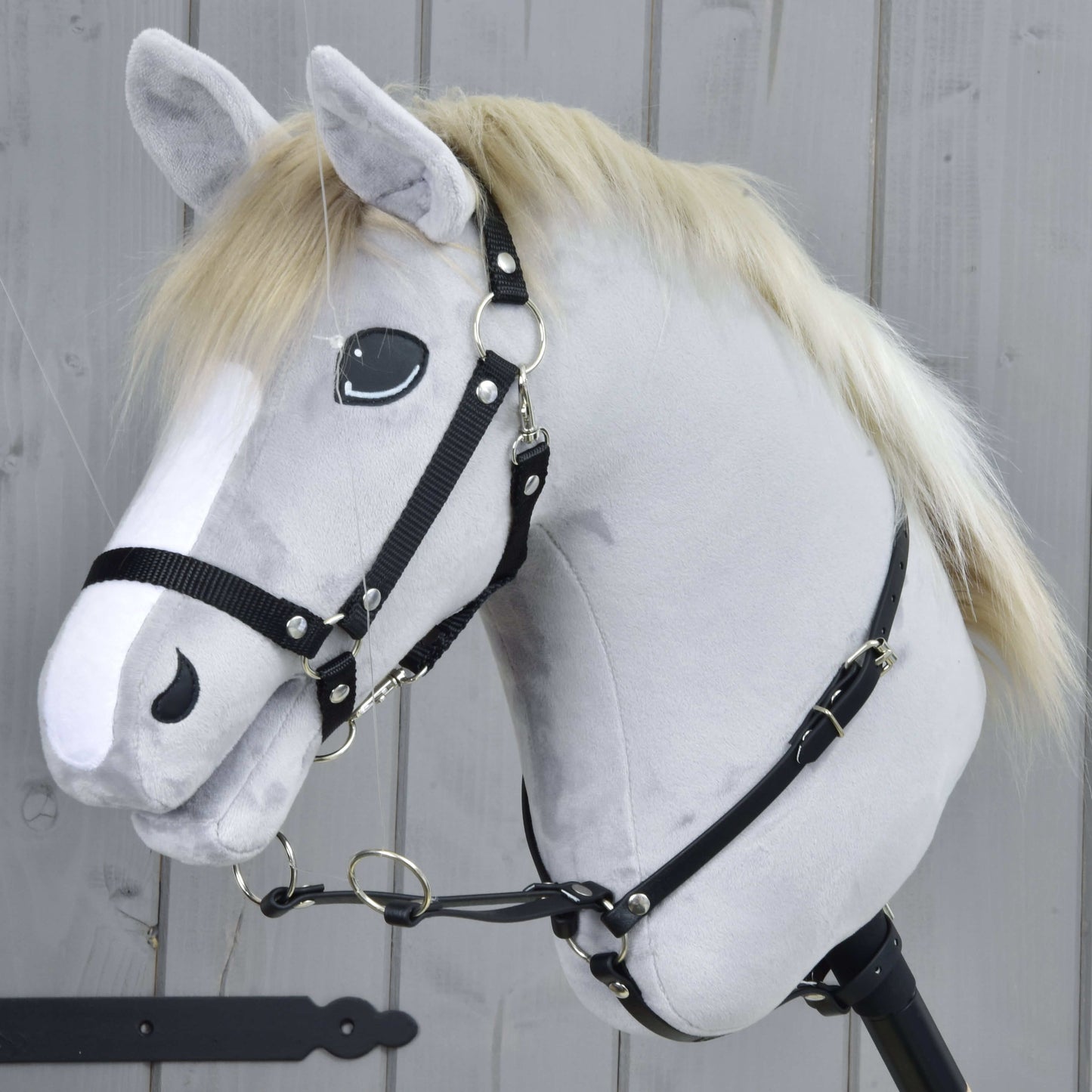 Martingal čierny od Hobby Horse LarDen na realistickej sivej hračke Hobby Horse so svetlou hrivou, bielou lysinou a čiernou uzdečkou, nasadenej na drevenej tyči pred sivým dreveným plotom.
