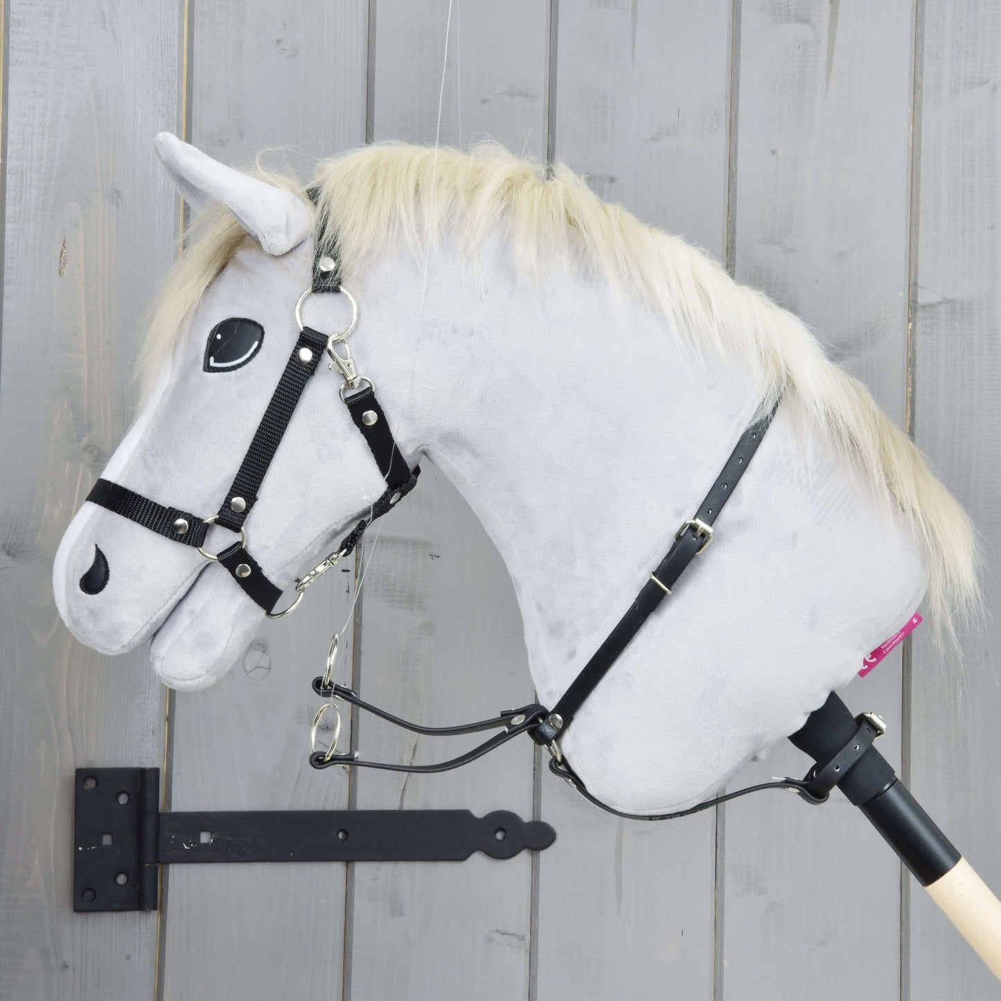 Martingal čierny od Hobby Horse LarDen na sivom plyšovom Hobby Horse s blond hrivou, čiernou uzdečkou, martingalom a opratami, nasadenom na drevenej tyči pred svetlosivou drevenou stenou.