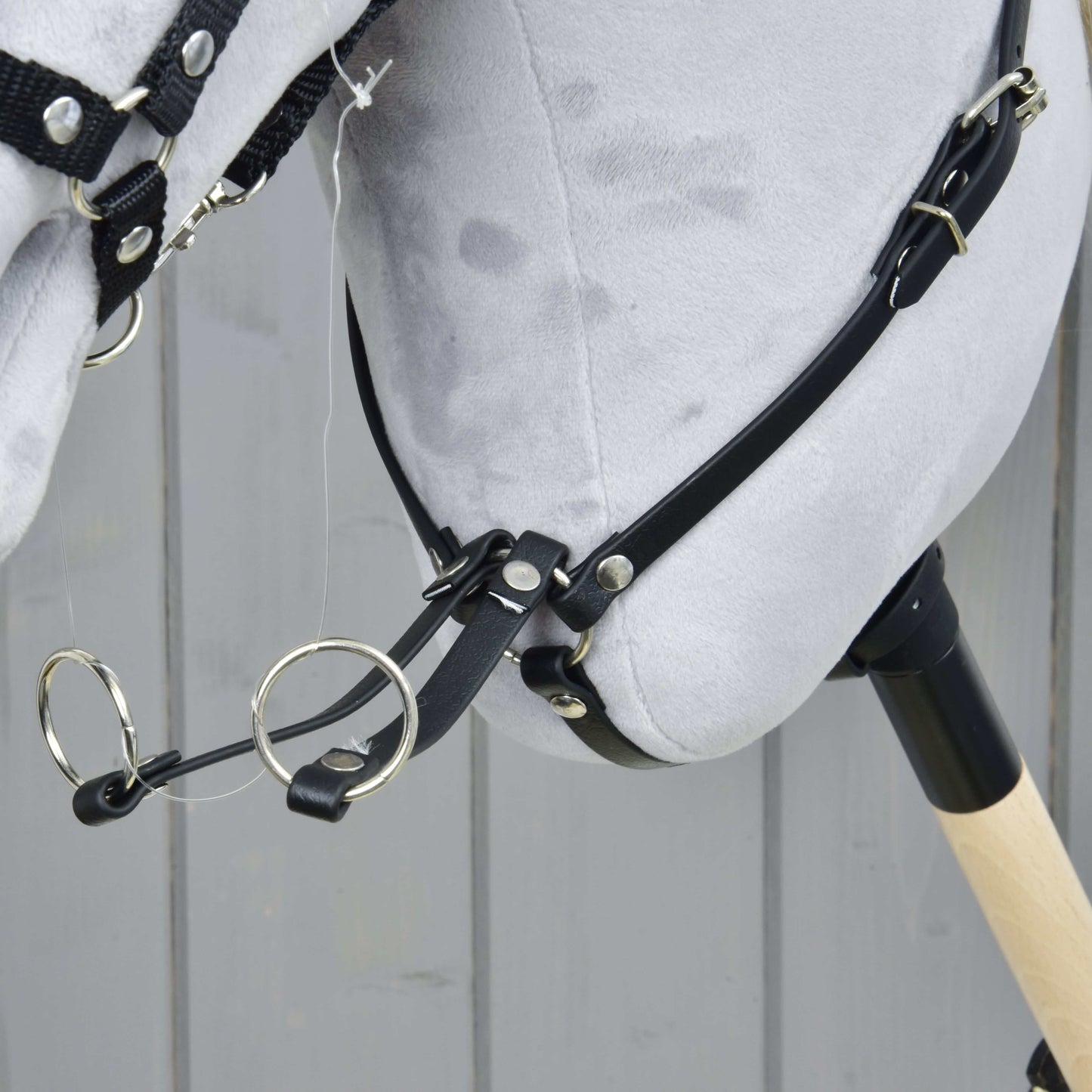 Detailný záber hlavy plyšového Hobby Horse Martingal čierny od Hobby Horse LarDen s čiernou uzdečkou, koženkovým martingalom a kovovými krúžkami na drevenej tyči, na pozadí sivých drevených panelov.