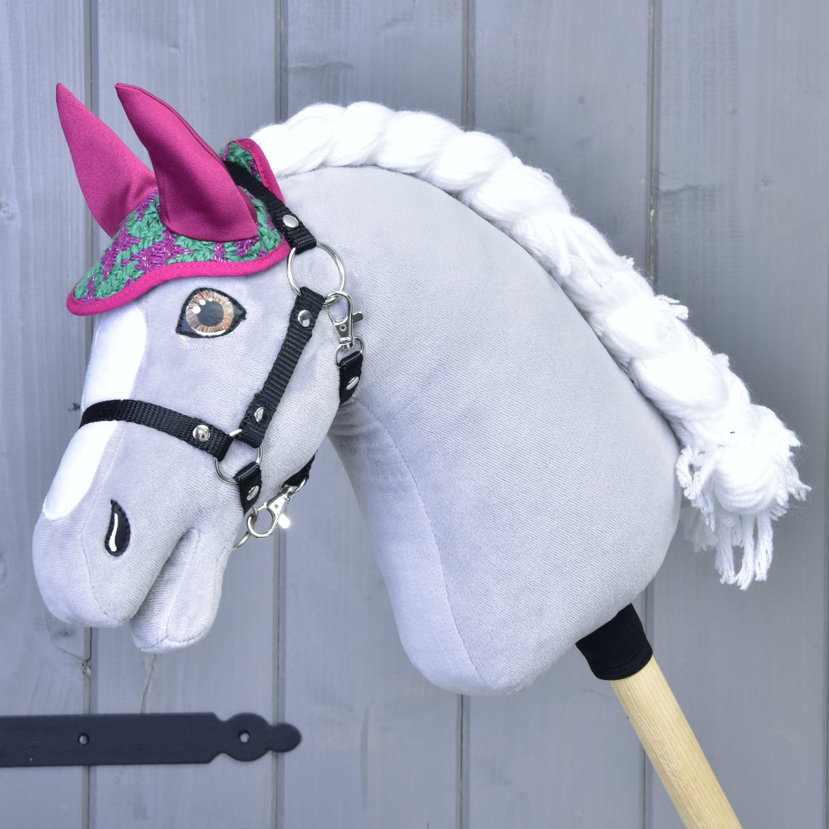 Set Coco od Hobby Horse LarDen so sivým telom, bielou hrivou, čiernou uzdečkou a ružovou čabrakou na uši stojí pred sivým dreveným pozadím.