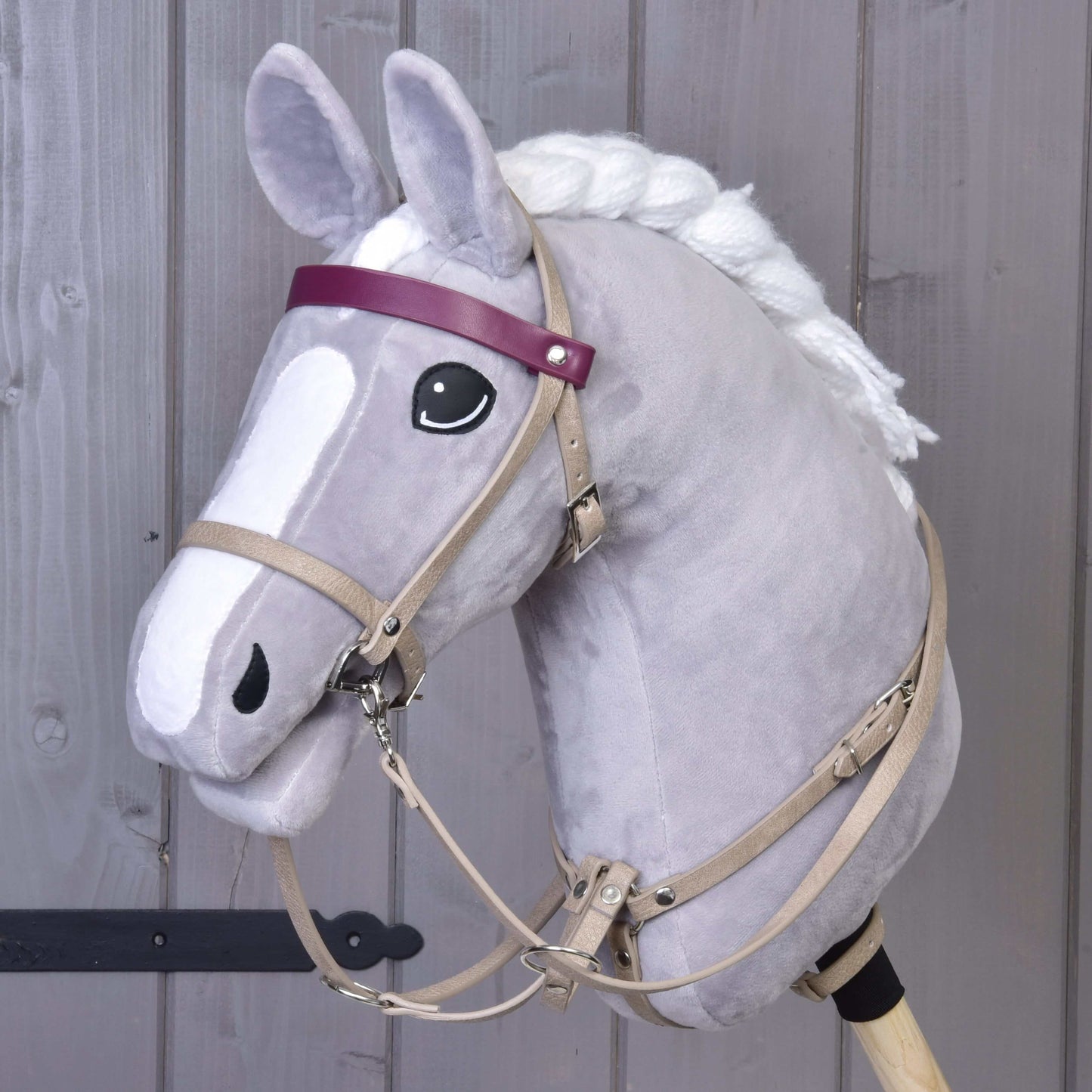 Sivý plyšový Hobby Horse od Hobby Horse LarDen s koženkovým setom Bordeaux – uzdečka, opraty a martingal –, bielou hrivou, čierno-bielymi očami a fialovou čelenkou stojí pred dreveným pozadím.