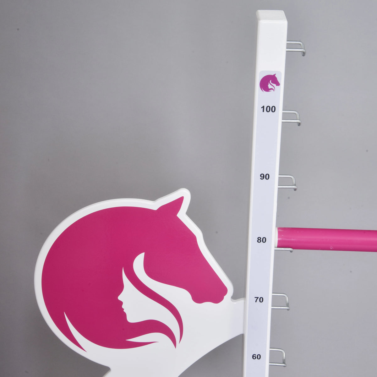 Prekážka LarDen kolmák 110 cm od Hobby Horse LarDen je biely stojan s nastaviteľnými značkami (60–100), kovovými háčikmi a ružovou konskou hlavou spojenou so siluetou ženy vľavo, na jednofarebnom sivom pozadí — ideálna pre jazdu na Hobby Horse.