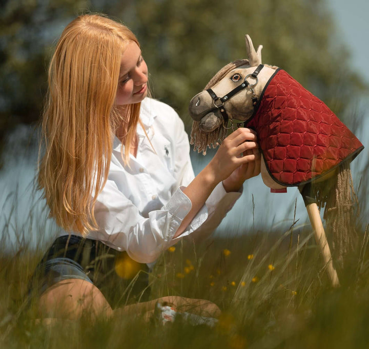 Mladý človek s dlhými blond vlasmi a v bielom tričku sedí vo vysokej tráve a jemne upravuje uzdečku Hobby Horse s červeným prešívaným prehozom, na pozadí rozmazanej zelene, a vystihuje nápaditého ducha Hobby Horsingu.