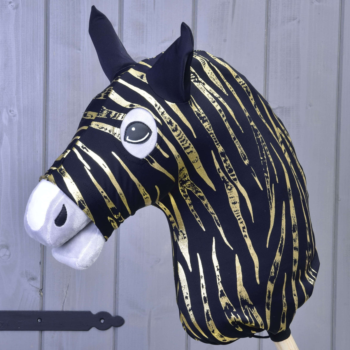 Hobby Horse LarDen ochranný oblek Zebra Gold (Lycra) s čierno-zlatou pruhovanou hlavou zebry, čiernymi ušami, bielym plsteným ňufákom a veľkými kreslenými očami, zobrazený pred svetlosivou drevenou stenou.