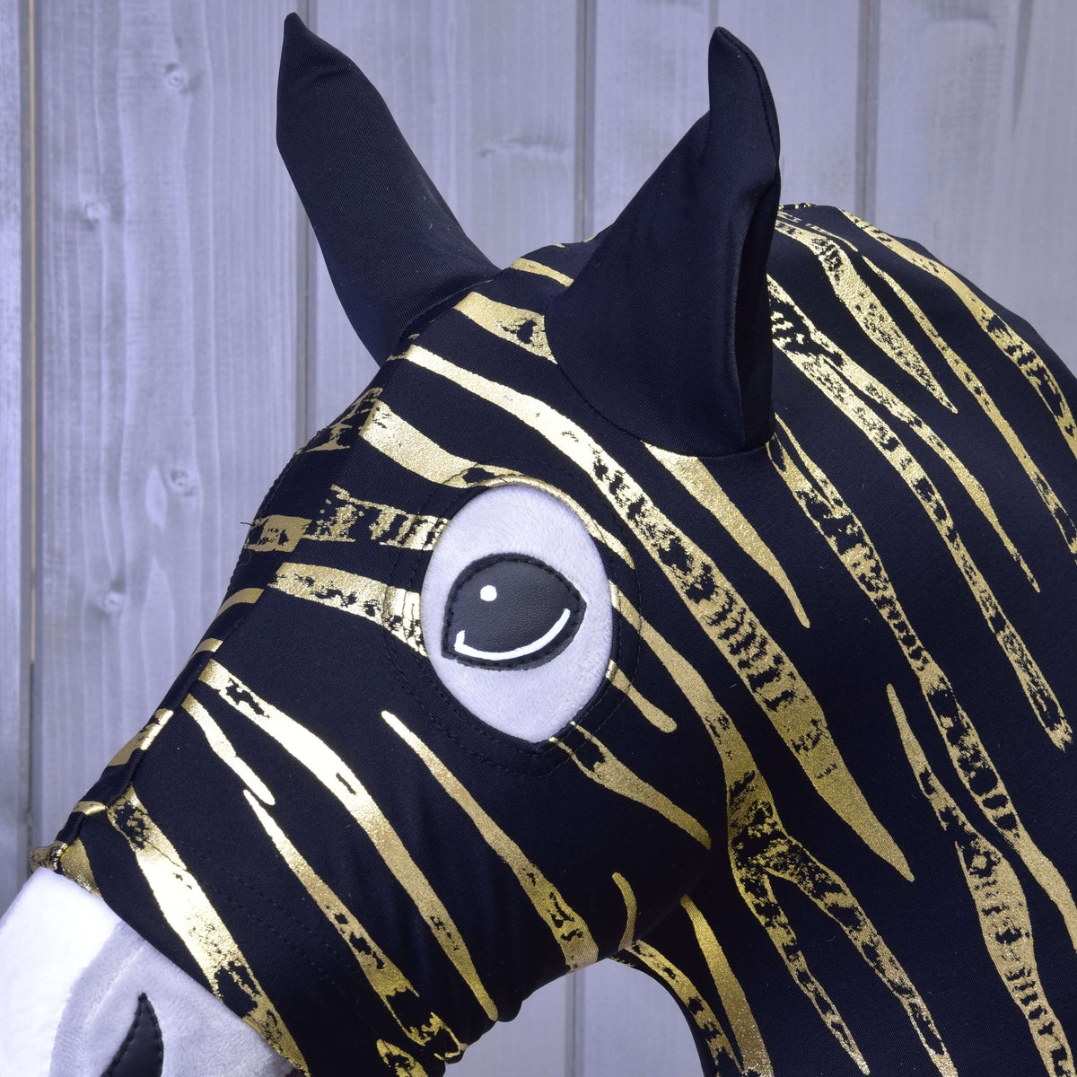 Detail ochranného obleku Hobby Horse LarDen Zebra Gold (Lycra) ukazuje čierno-zlato pruhovanú látku, veľké čierne uši a kreslené oko na pozadí svetlého dreva.