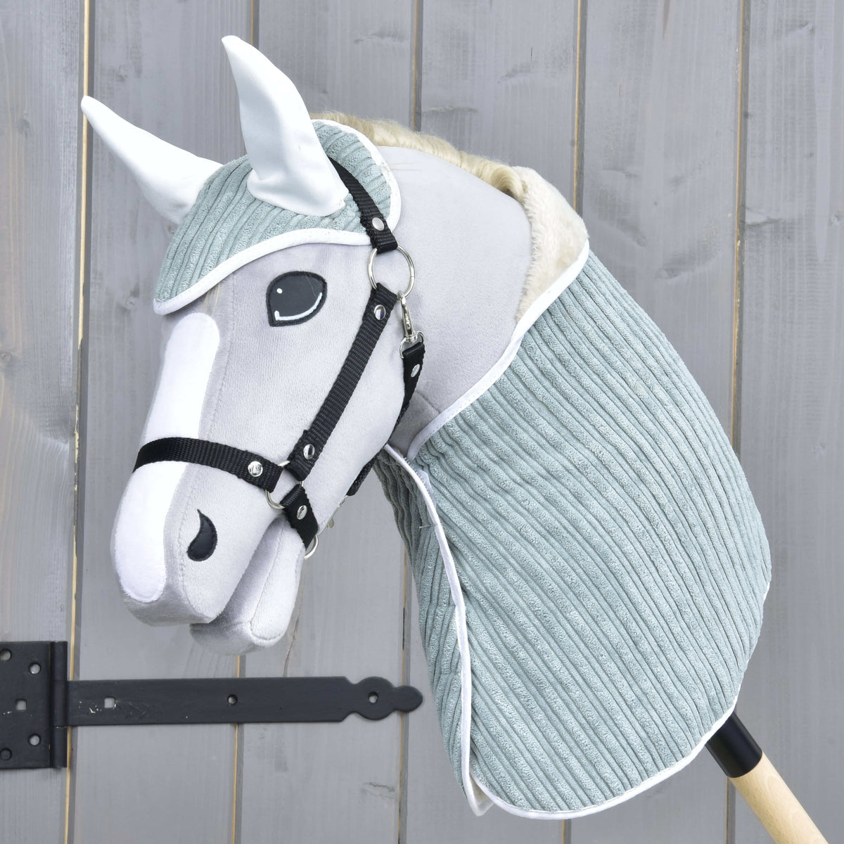 Set Manchester Little Green od Hobby Horse LarDen so sivo-bielym Hobby Horse s mäkkou látkovou hlavou, čiernou uzdečkou, čabrakou a svetlým modrozeleným krytom krku na drevenej tyči, zobrazený pred dreveným plotom.