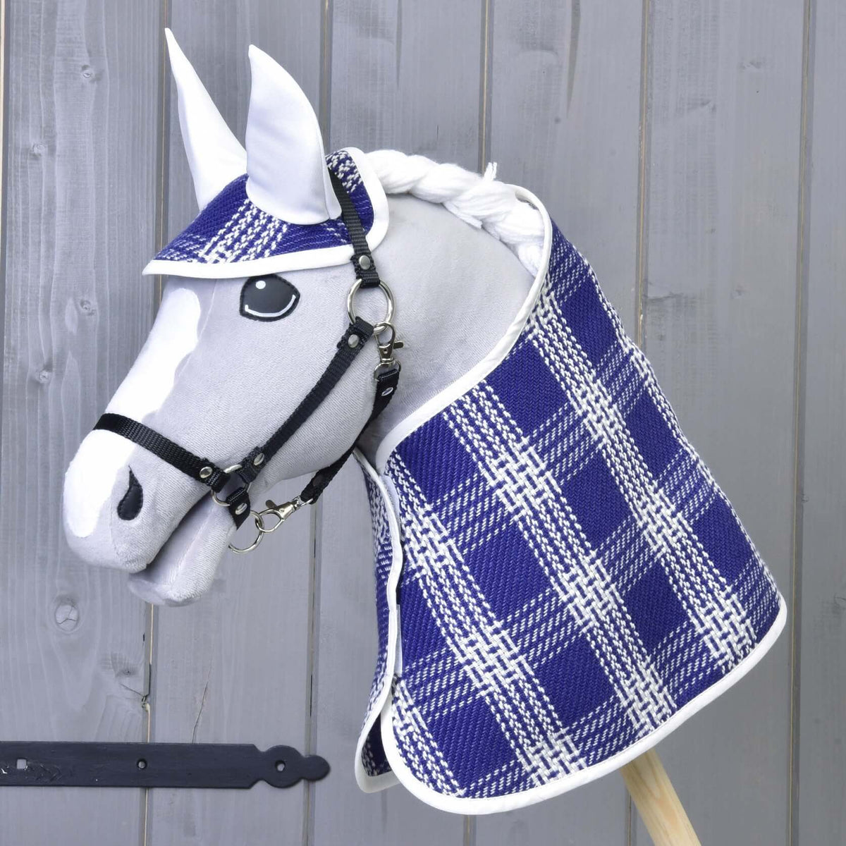 Biela hračka Hobby Horse LarDen s čiernou uzdečkou má na sebe károvanú deku a čiapku Set Blue Cube a stojí pred sivým dreveným pozadím.