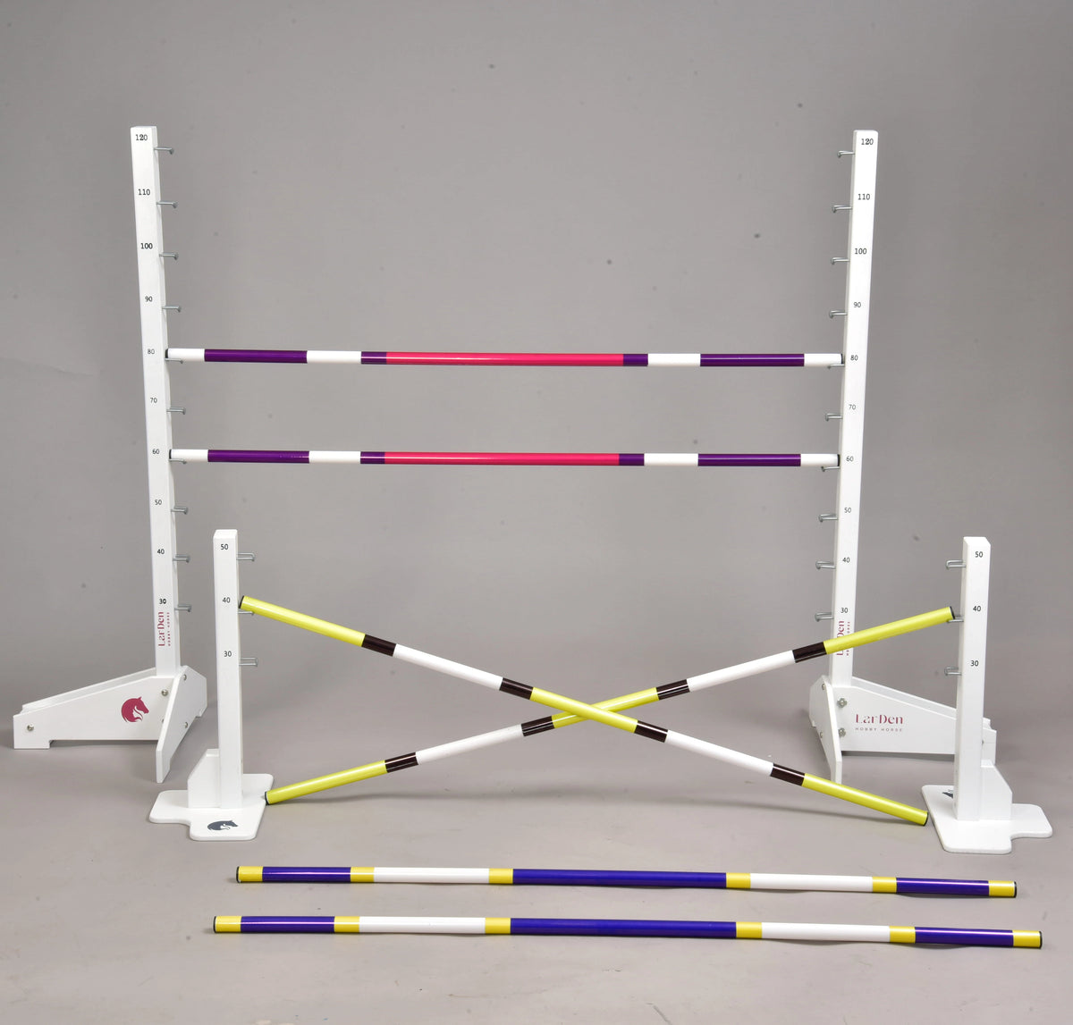 Set prekážok pre Hobby Horsing od Hobby Horse LarDen s nastaviteľnými bielymi stojanmi s meracími značkami a farebnými tyčami vo fialovej, ružovej, žltej a čiernej farbe – ideálny na skoky alebo cavaletti, niektoré tyče sú postavené a iné ležia na podlahe.