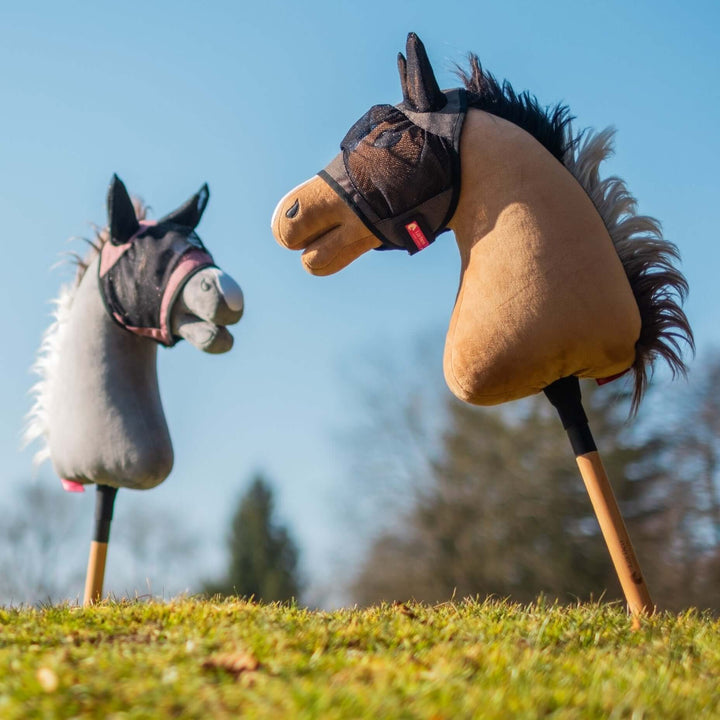 Dva hračkárske Hobby Horse s plyšovými hlavami, jeden sivý a jeden hnedý, stoja vzpriamene na trávnatej zemi proti jasne modrej oblohe a rozmazaným stromom. Oba kone majú malé látkové uzdečky, pripravené na nápadité dobrodružstvo v Hobby Horsingu.